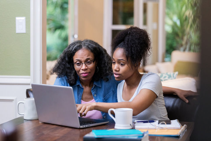 Young woman and her mature mom look at something on a laptop as the young woman studies for a college exam.