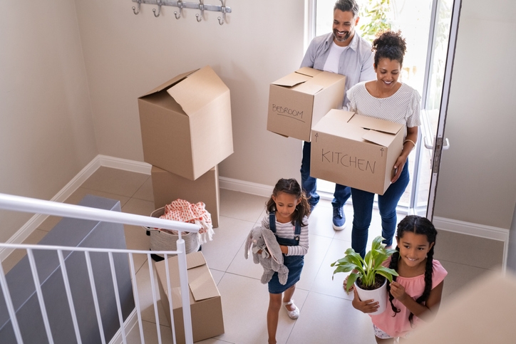 family with two children carrying boxes and plant in new home on moving day. High angle view of happy smiling daughters helping mother and father with cardboard boxes in new house. Top view of excited kids having fun walking up stairs running to their rooms while parents holding boxes.
