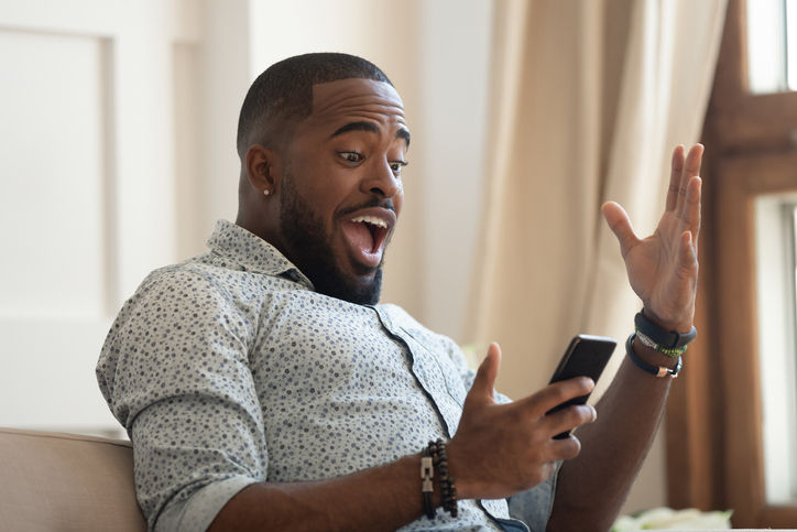 Surprised happy african man holding phone looking at cellphone read good news in sms sit on sofa, amazed black guy winner excited by mobile app win scream with joy celebrate victory success at home