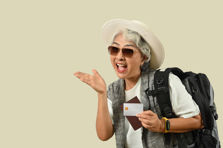 Smiling mature female traveler with backpack holding credit card and pointing at copy space, ready for summer vacation, journey trip.