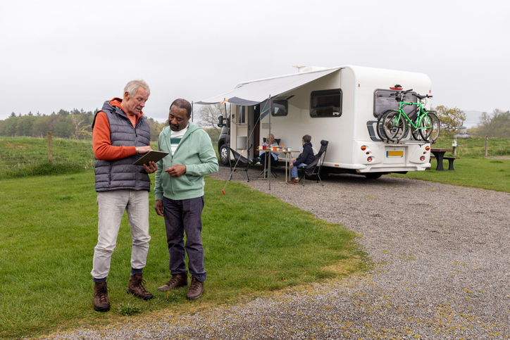 Group of senior friends on a hiking staycation, staying in a camper van in Dumfries and Galloway, Scotland. Two men are standing looking at a digital tablet together while women sit behind them in front of a camper van.