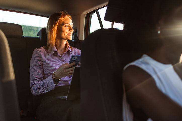 Photo of an unrecognizable African-American woman driving a car, while a modern mature woman is sitting in the back seat, using a laptop and a mobile phone