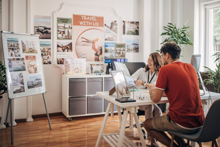 Two people, man in modern travel agency talking with woman who is working there, she is showing him of places for vacation.