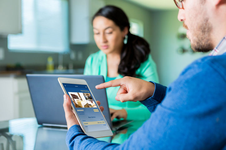 Man uses digital tablet to make hotel reservation in the comfort of his home. His wife is in the background using a laptop.