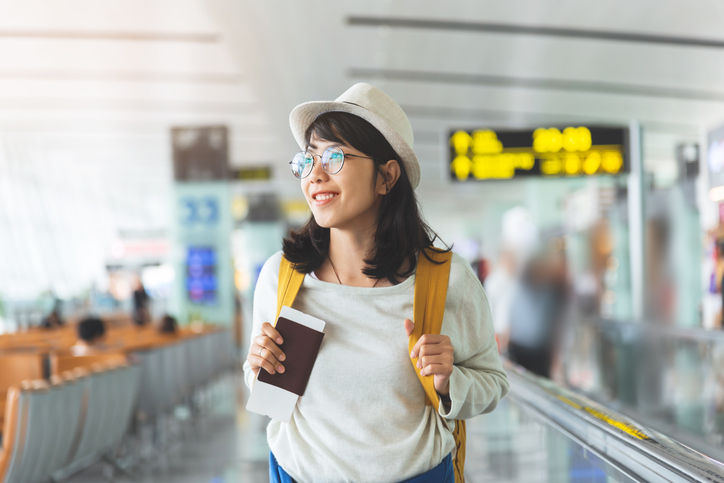 Happy Asian woman wear glasses, hat with yellow backpack is holding flying ticket, passport while waiting for the flight at the hall of airport. Smile.