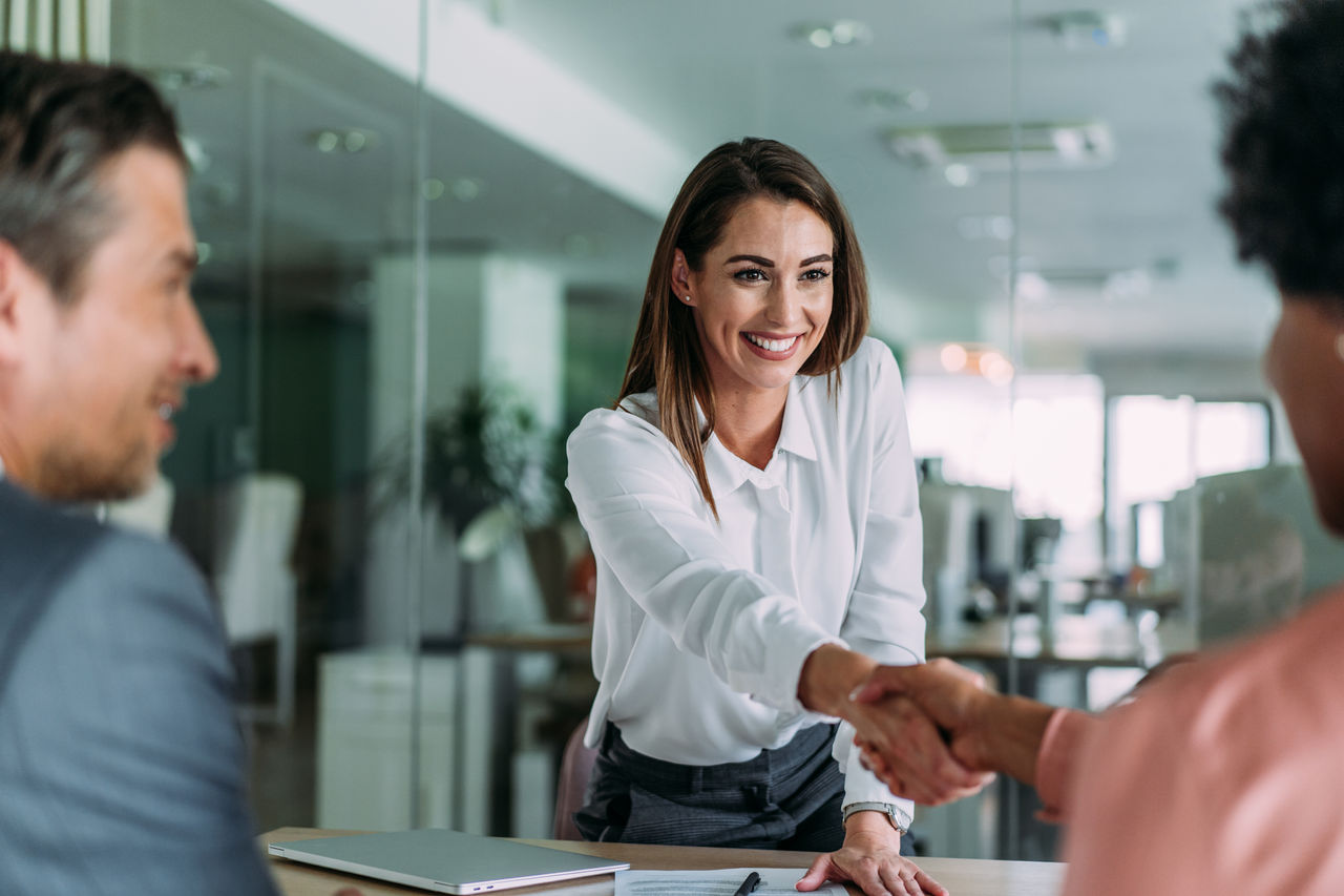 Businesspeople handshaking across the table during a meeting in modern office. Group of business persons in business meeting. Shot of three entrepreneurs on meeting in board room. Corporate business team on meeting in the office.