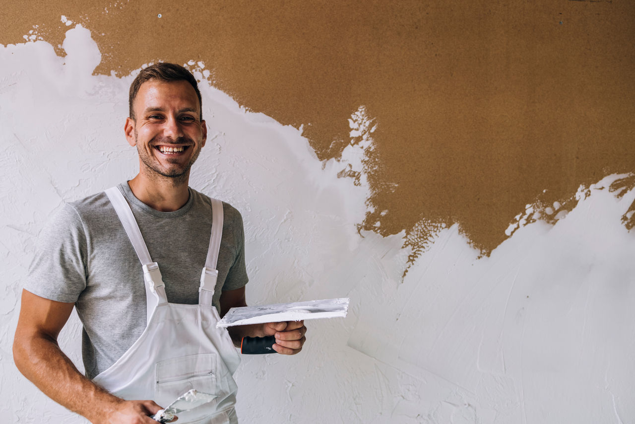 Cheerful man looking at camera while fixing wall surface and preparing wall for painting at home