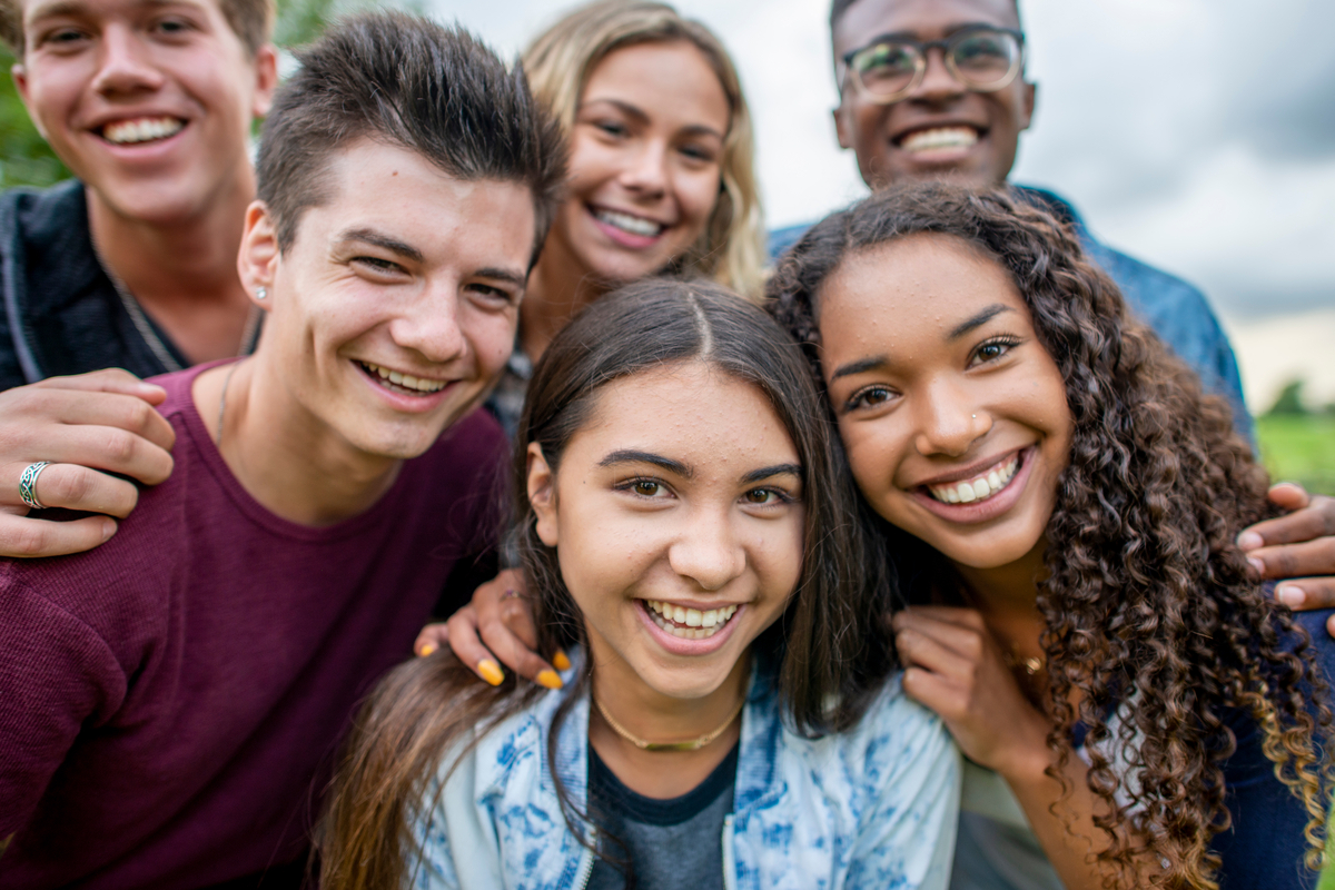 A multi-ethnic group of high school students are outdoors on a summer day. They are gathered together to take a selfie, and they are all smiling.