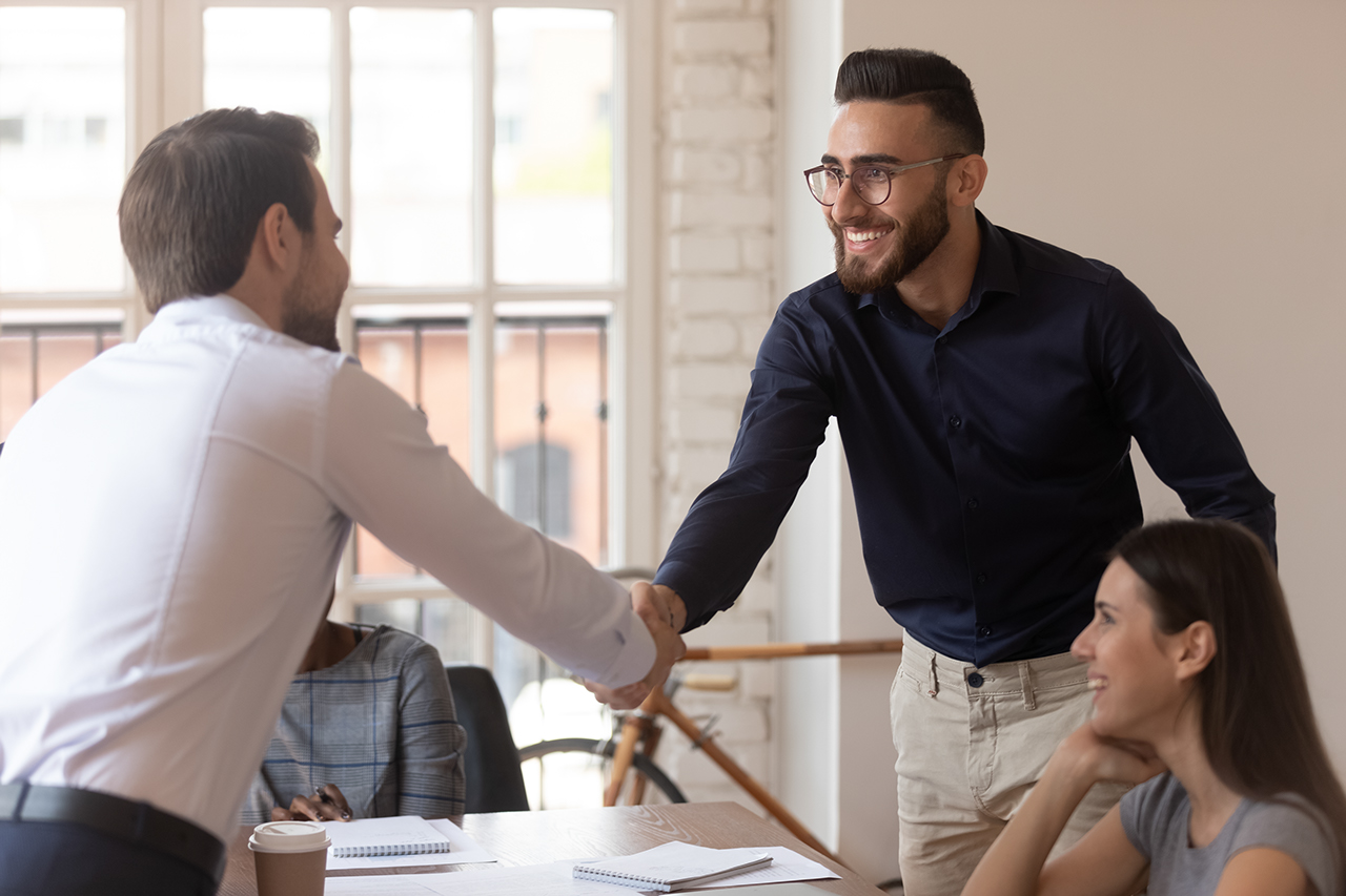 Confident male team leader shaking hands with young smiling arabic employee, welcoming new colleague at team brainstorming meeting at office. Two happy multiracial businessmen making agreement.