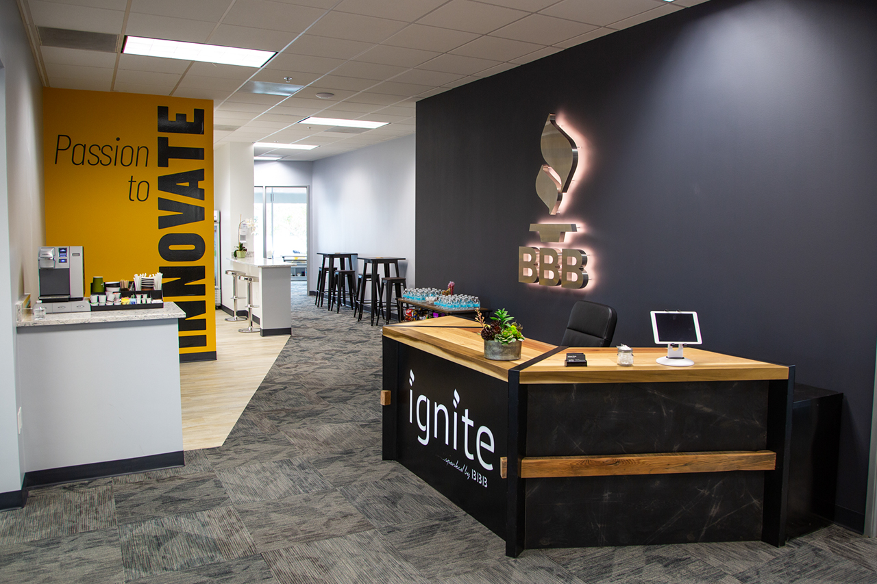 black industrial metal and wood front desk reading ignite sparked by BBB with BBB logo on the wall behind. Passion to innovate words on a yellow wall. Cafe table and kitchen in the background.