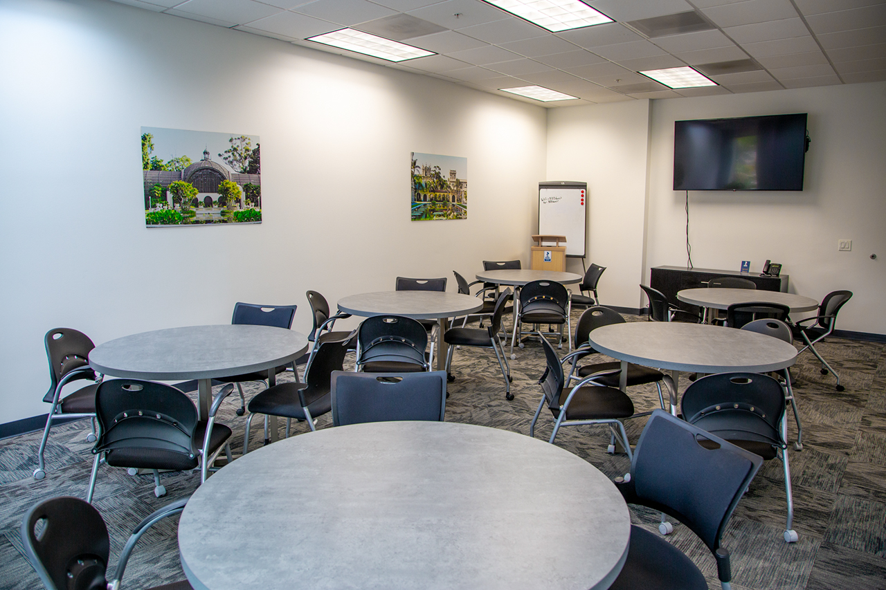 large room with many round gray tables and a tv screen on the wall at the front of the room