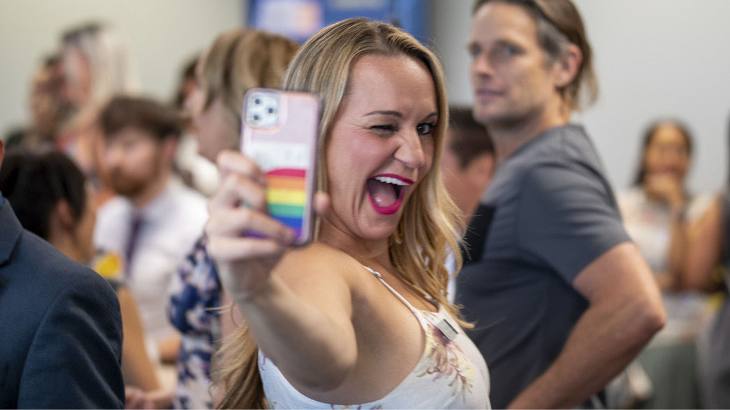 Smiling blonde female taking selfie with cellphone at a crowded networking event