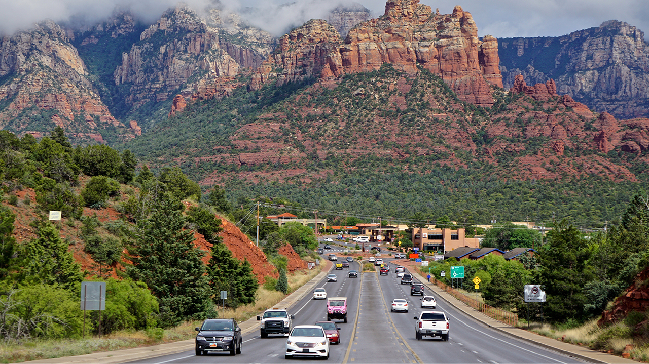 View of Sedona, Arizona with trees, a shot of the mountain and a roadway with multiple cars leading into town